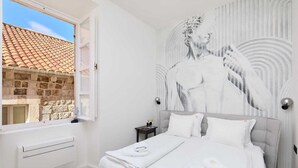 Un dormitorio con una cama grande, ropa de cama blanca, una mesita de noche, una lámpara de pared y vistas a un tejado con tejas a través de una ventana.