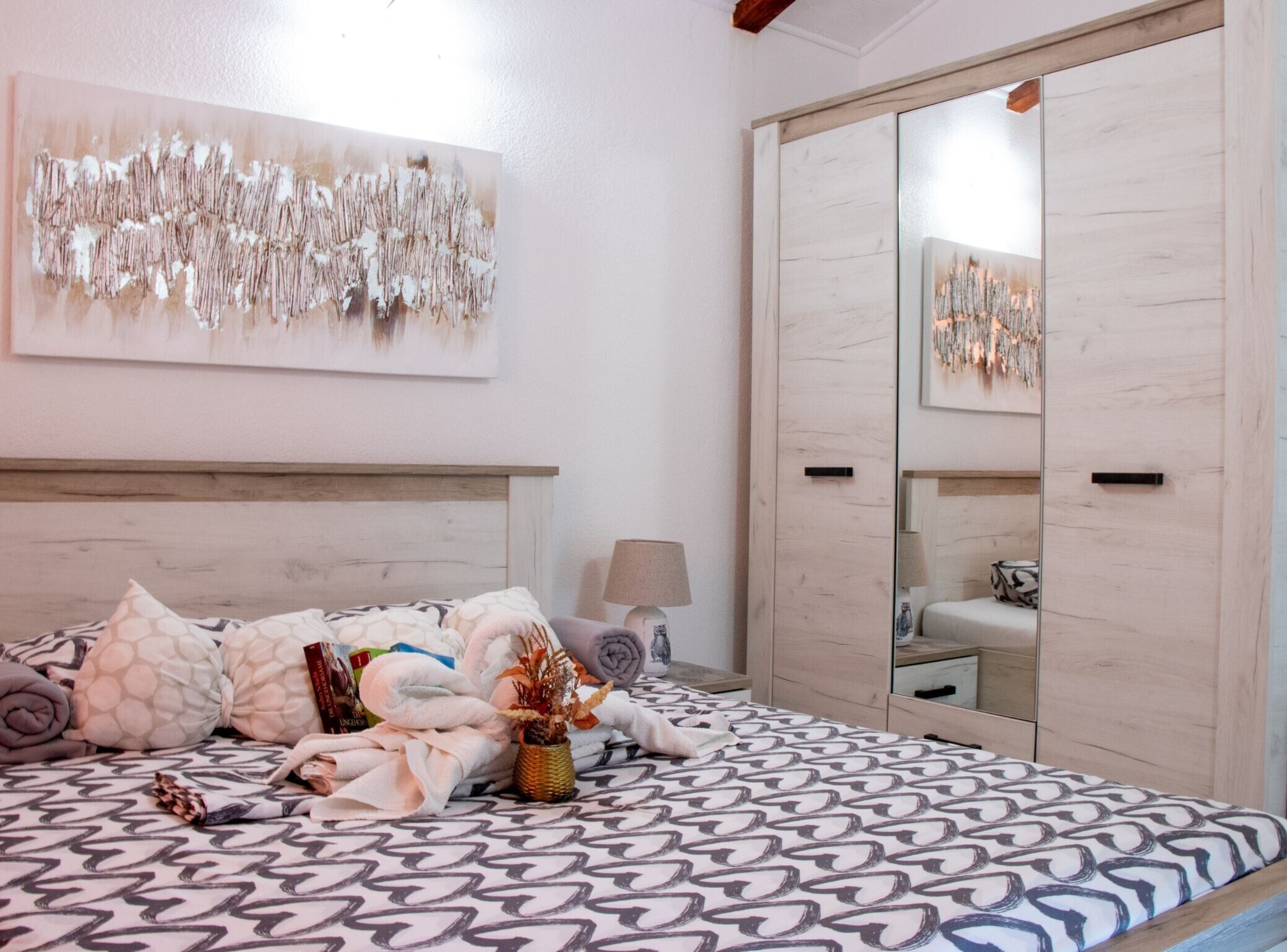 A bedroom with a patterned bedspread, a headboard, a bedside lamp, a mirror, and a wall-mounted abstract painting.