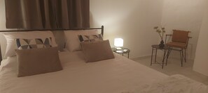 Een net opgemaakt bed met meerdere kussens, een nachtkastje met een lamp, en een stoel in een kamer met effen wanden.
