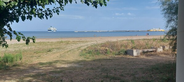 Een strand met een cruiseschip in de verte, een pier met boten, en enkele mensen op het zand.