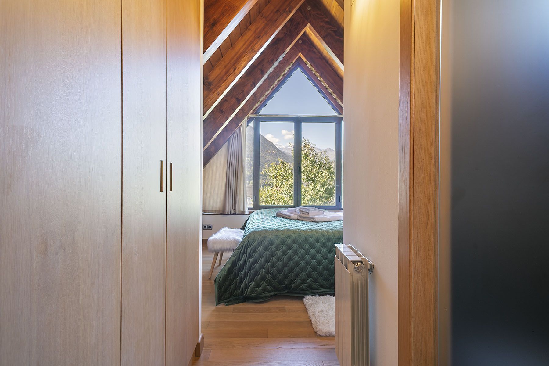 A narrow hallway leading to a bedroom with a large window, wooden beams, and a bed with a green quilt.