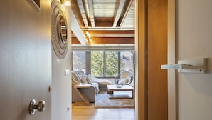 A room with a wooden ceiling, a bed with a white blanket, a wicker chair, and a view of trees through large windows. - Luderna - Dúplex Sant Marti XII (Gessa)