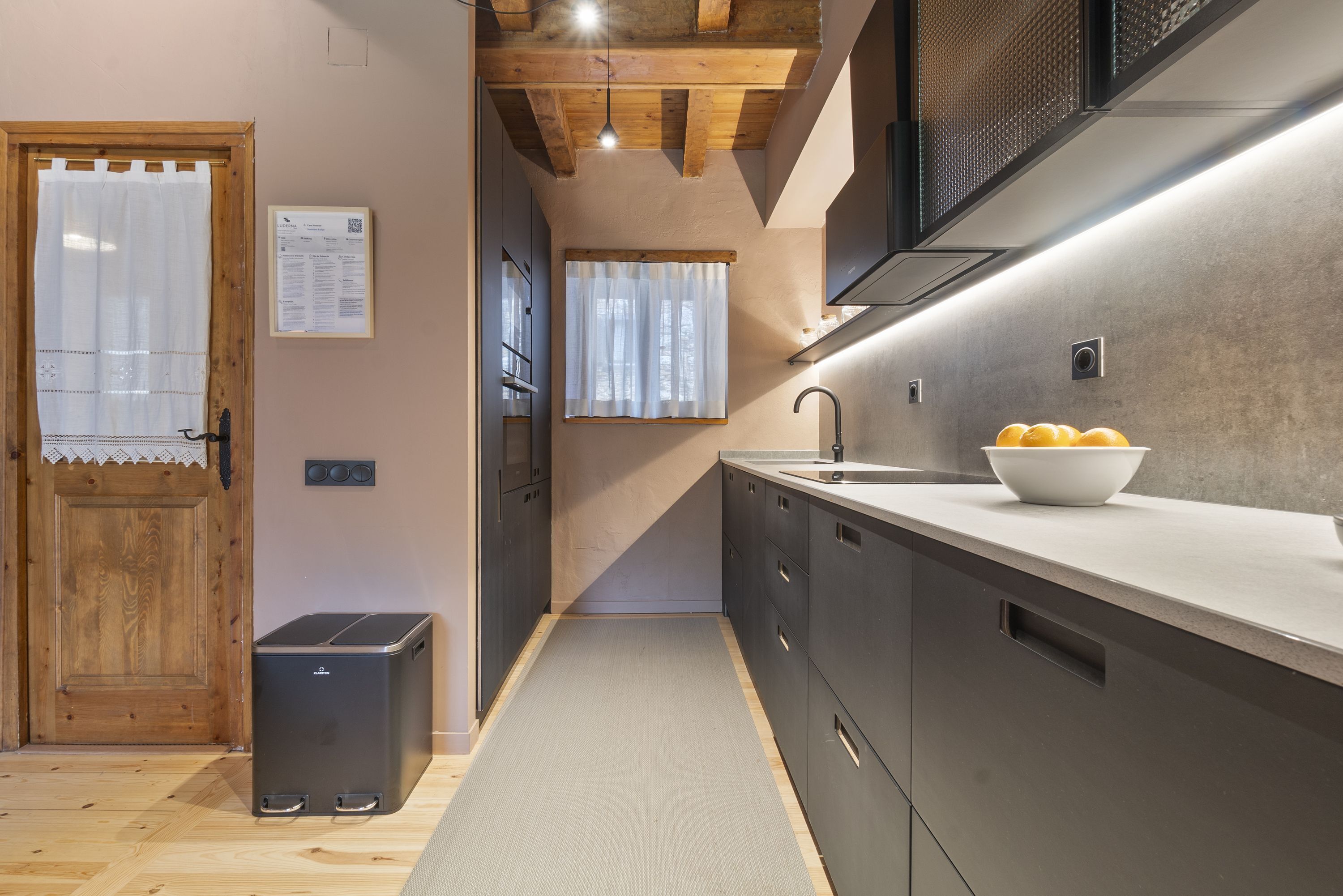 A narrow kitchen with wooden beams, a sink, cabinets, and a bowl of fruit on the counter.