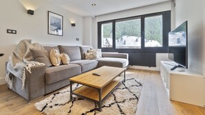 A modern living room with a grey sofa, a wooden coffee table, and a large window with a view of buildings and trees. - Luderna - Apartamento Misharnon (Baqueira)