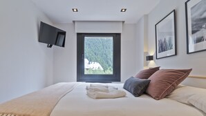 A bedroom with a bed, a TV mounted on the wall, a window with a view of trees, and framed pictures on the wall. - Luderna - Apartamento Misharnon (Baqueira)