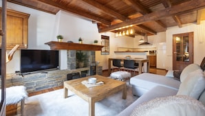 A cozy living room with a stone fireplace, wooden ceiling beams, a flat-screen TV, and a comfortable seating area. - Luderna - Apto con terraza Rouche (Arties)