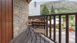 A wooden deck with a stone wall, a table with a plate of food and drinks, and a view of a mountainous landscape. - Luderna - Apto con terraza Rouche (Arties)