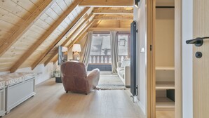 Ein Schlafzimmer im Dachgeschoss mit hohen Decken, Holzbalken, einem braunen Ledersessel, einem Nachttisch mit Lampe und Blick durch ein Fenster auf einen Balkon.