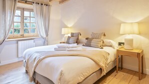 Een net opgemaakt bed met witte lakens en een beige deken, een nachtkastje met een lamp, en een raam met gordijnen.