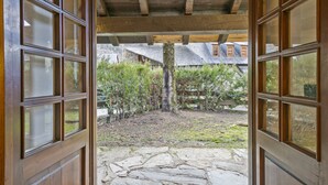 Een open houten deur die leidt naar een met stenen bestraat binnenhof met een houten plafond en uitzicht op een huis op de achtergrond.