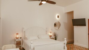 Un dormitorio con cama blanca, ventilador de techo, lámparas de noche, televisor en la pared y piso estampado.