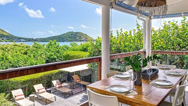Un coin repas avec une table dressée pour quatre personnes, donnant sur un jardin luxuriant et offrant une vue panoramique sur des collines et un plan d’eau.