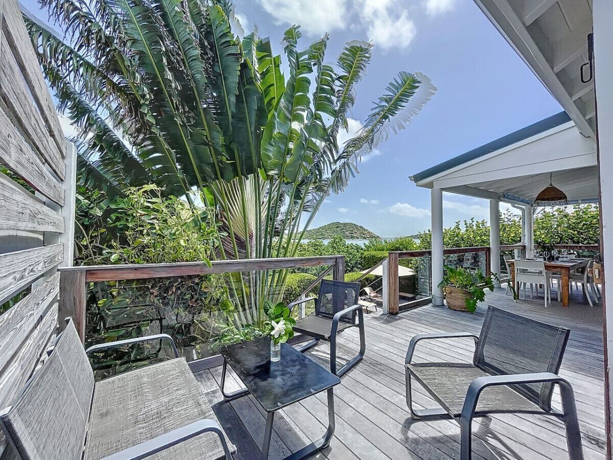 A wooden deck with outdoor furniture, including chairs and a table, surrounded by lush greenery and a view of the landscape.