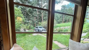 A room with a large window overlooking a grassy area and trees. There is a bed with a gray mattress and two pillows.