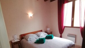 Ein Schlafzimmer mit Holzboden, einem Bett mit weißer Bettwäsche und grünen Handtüchern, einem Nachttisch mit Lampe sowie einem Fenster mit roten Vorhängen.