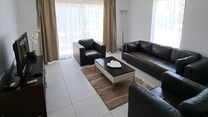 Una sala de estar moderna con dos sofás de cuero negro, una mesa de centro, un televisor sobre un mueble bajo y un ventanal con cortinas.