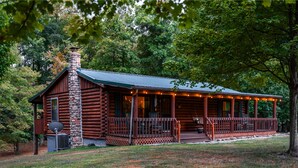 A log cabin with a stone chimney, a porch with chairs, and a covered deck area. - Havenbrook~Rustic luxury, 10acres, hot tub (New Plymouth)