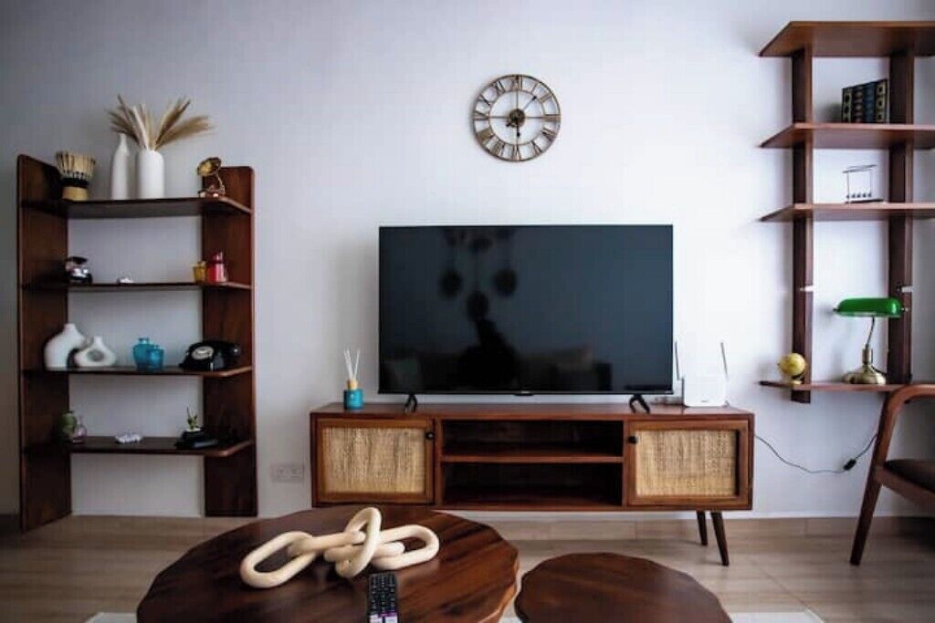 Una sala de estar con un televisor sobre un mueble de madera, un reloj en la pared y estantes con diversos objetos.