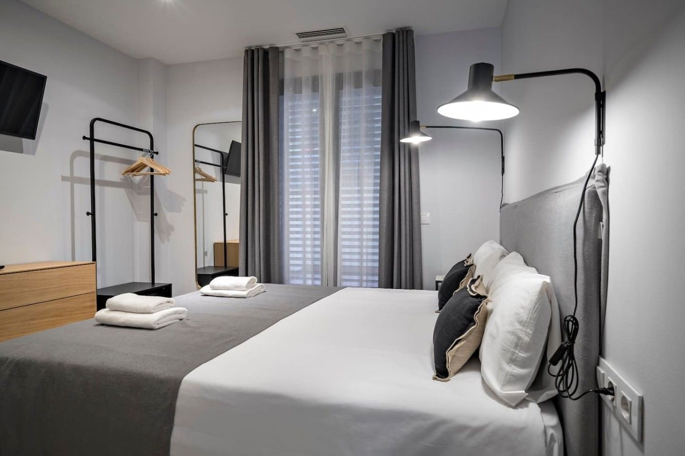 A modern hotel room with a bed, bedside lamp, and a view of the city through large windows.