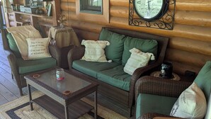Interior acogedor de una cabaña de troncos con un sofá verde, sillas de mimbre, una mesa de centro y un reloj de pared.