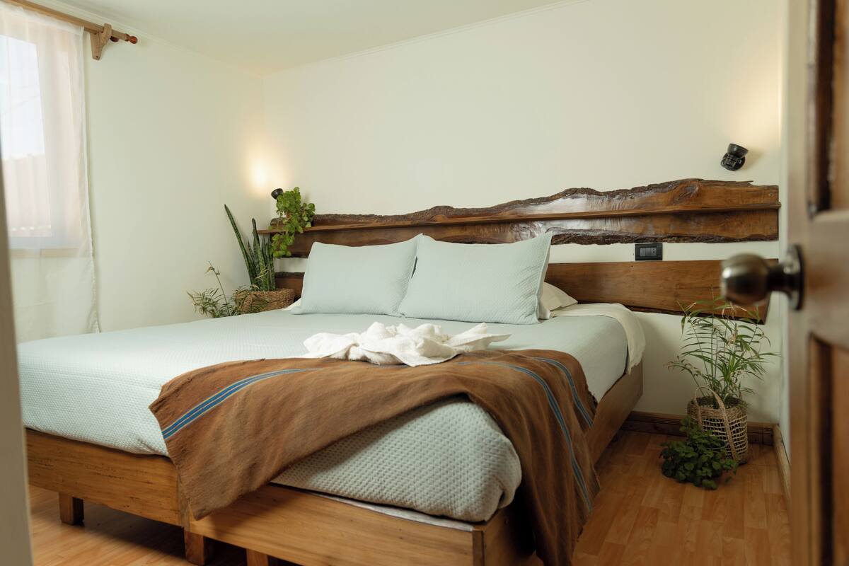 A neatly made bed with a wooden headboard, a white bedspread, and a brown throw blanket. There is a potted plant on the floor beside the bed.