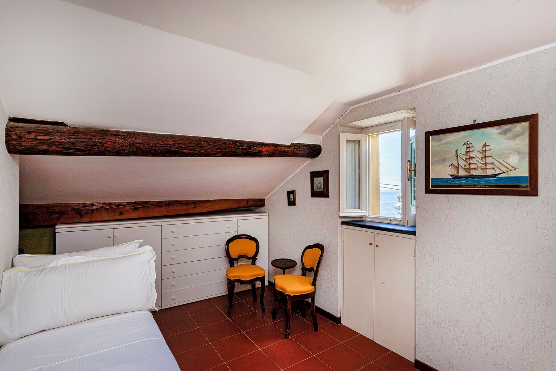 A small room with a bed, two chairs, a small table, and a window with a view of the sea.