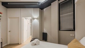 Une chambre moderne avec un lit, un radiateur, une fenêtre avec des stores et un couloir menant à une autre pièce.