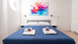 Een hotelkamer met een groot bed, twee kussens, een blauw geborduurd beddegoed, een kleurrijk abstract schilderij aan de muur en een witte deur.