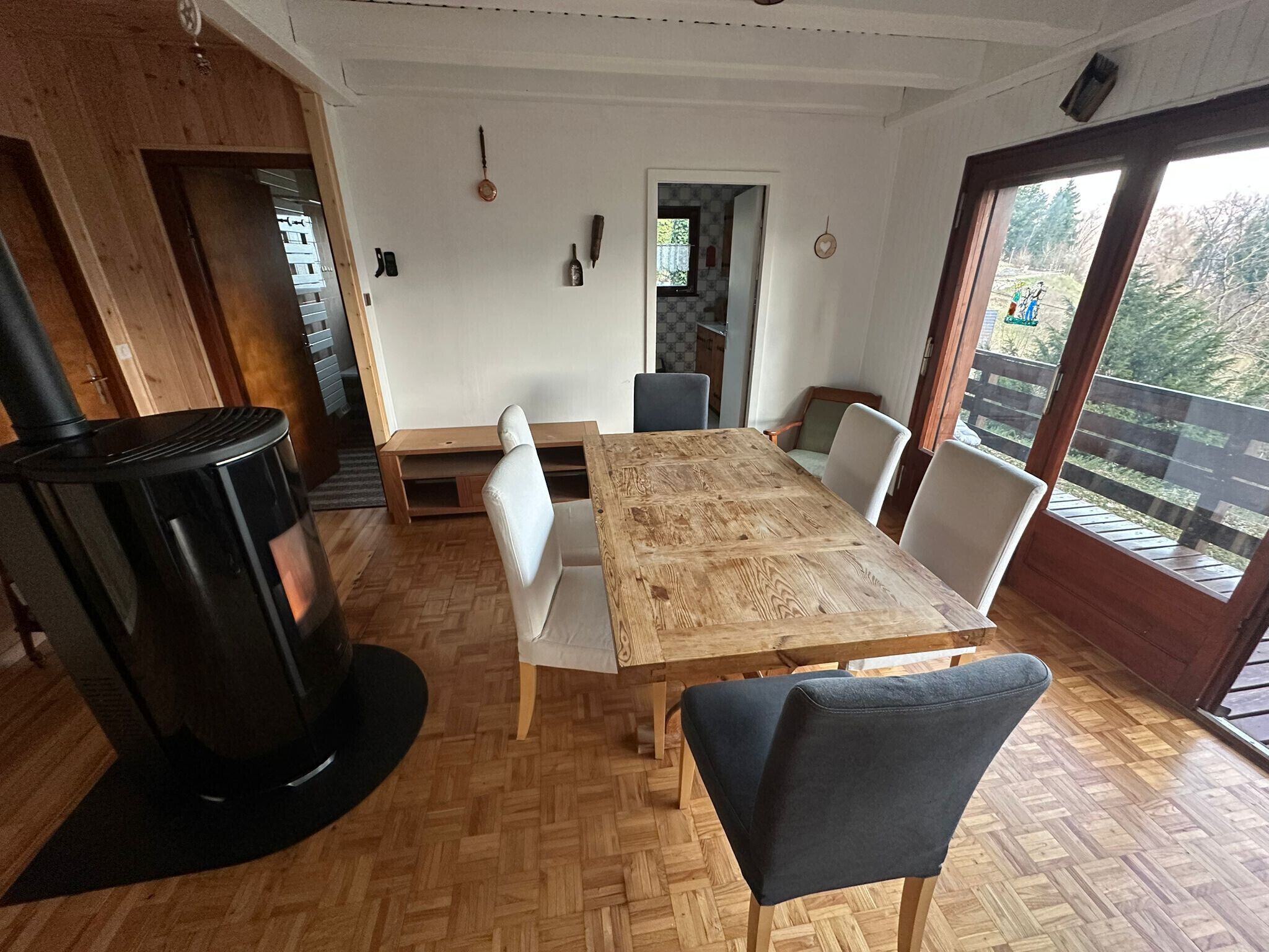 Un coin repas avec une table en bois, des chaises, un poêle noir et une vue à travers une porte vitrée.