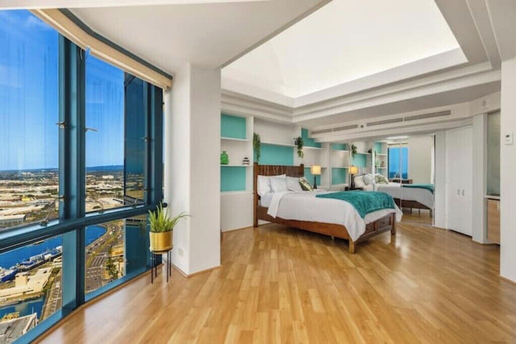 A spacious bedroom with a large bed, a view of the city through floor-to-ceiling windows, and a skylight.