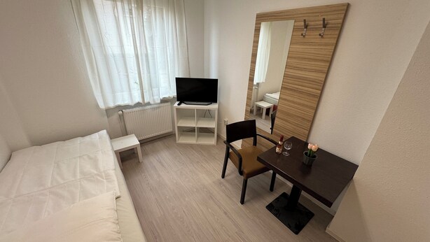 Hotellihuone, jossa on sänky, pieni työpöytä tuolilla, jalustalla oleva televisio, peili ja ikkuna, jossa on verhot.