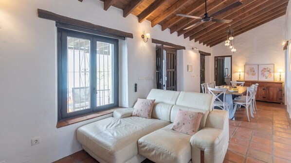 A spacious living area with a large sofa, dining table, and chairs. There are large windows with metal bars, a ceiling fan, and a kitchen area visible in the background. - Finca Mojito Cómpeta by Ruralidays (Málaga)