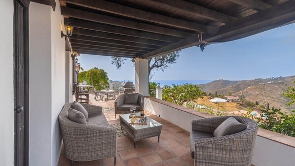 A covered outdoor seating area with wicker furniture, a table with a bottle and glasses, and a scenic view of hills and a body of water. - Finca Mojito Cómpeta by Ruralidays (Málaga)