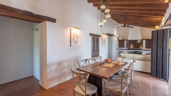 A dining area with a wooden table and chairs, a kitchen in the background, and a ceiling fan. - Finca Mojito Cómpeta by Ruralidays (Málaga)
