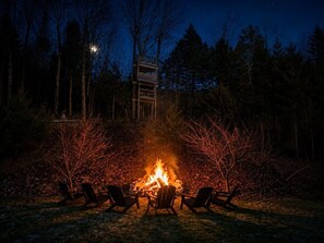 Ein Lagerfeuer mit Stühlen darum herum, umgeben von Bäumen und einer Holzhütte im Hintergrund.