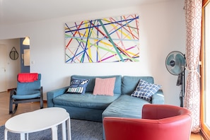 Un salon avec un canapé bleu, des fauteuils rouges et bleus, une table blanche et un tableau abstrait aux couleurs vives accroché au mur.