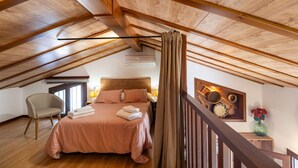 Un dormitorio con techo inclinado, vigas de madera, cama con ropa de cama rosa, una silla y una mesita de noche con una lámpara.