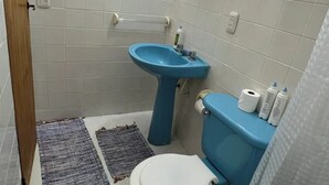 Un baño con lavamanos, inodoro y cortina de ducha azules. Hay un espejo en la pared y una pequeña ventana sobre el lavamanos.