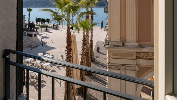 A balcony with a view of a beach, palm trees, and a building with classical architecture. - Apartment 'Flat 3' with Lake View, Wi-Fi and Air Conditioning (Salò)