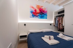 A modern bedroom with a blue quilted bedspread, a white pillow, a wall-mounted abstract painting, a wardrobe, and a bedside table.