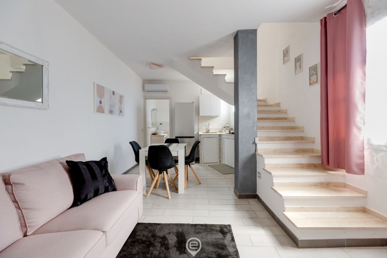 A modern living room with a sofa, a dining area with chairs, a staircase, and a kitchen in the background.