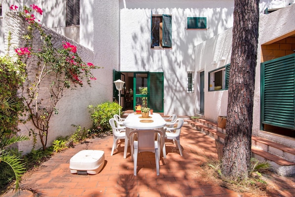Une cour avec une table et des chaises blanches, entourée d’un bâtiment blanc aux volets verts.