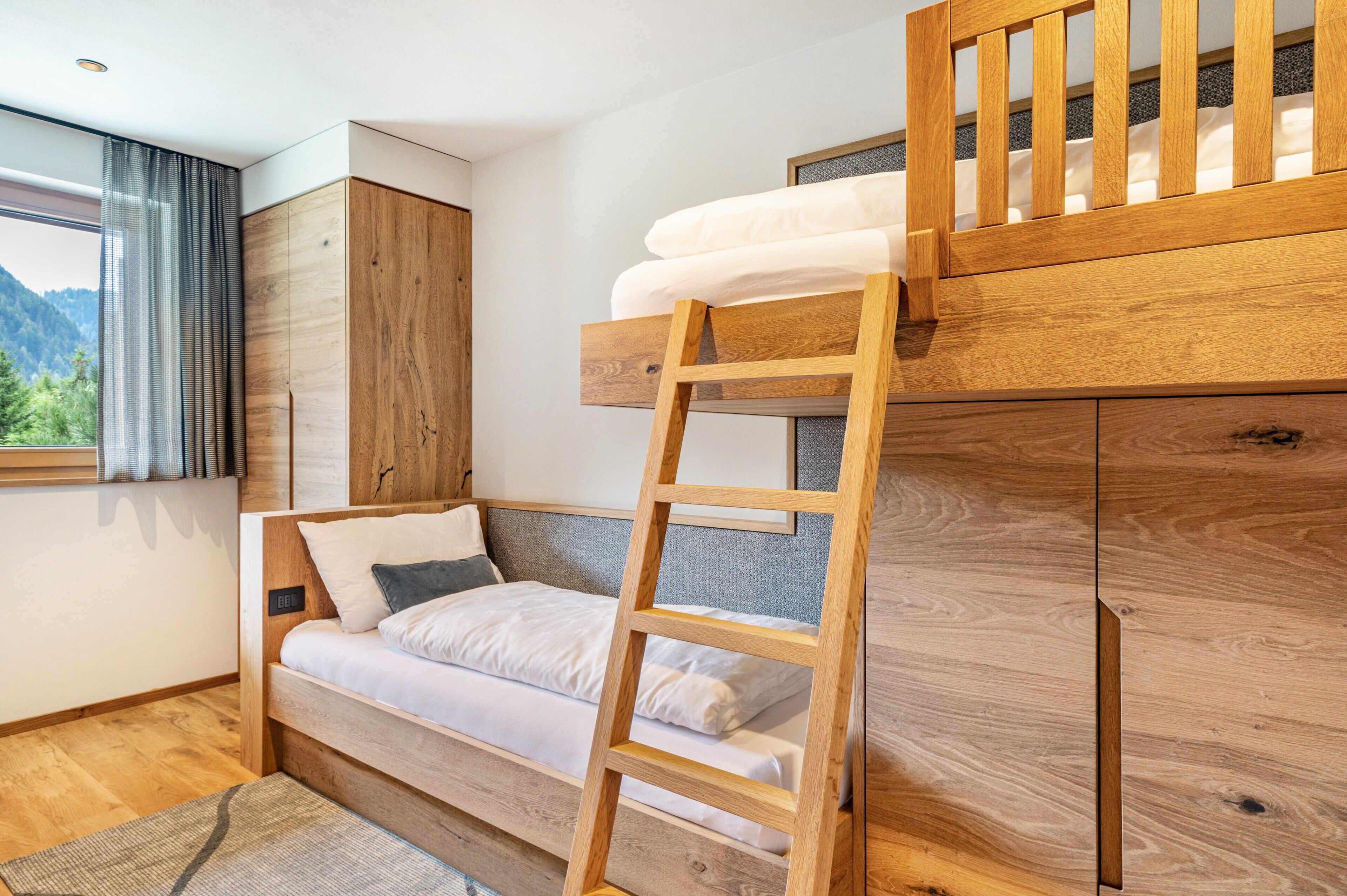 A bunk bed with a ladder, a wooden wardrobe, a window with curtains, and a bed with pillows.
