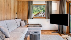 Ein modernes Wohnzimmer mit einem Ecksofa, einem Flachbildfernseher, Holzboden und einem Fenster mit Vorhängen.