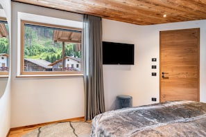 Een slaapkamer met een groot raam, een flatscreen televisie, een houten deur en een klein krukje.