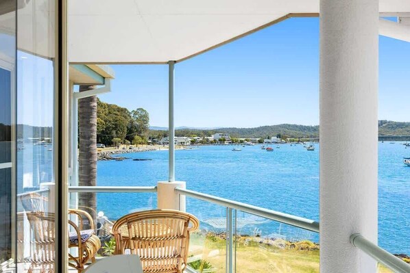 A balcony with wicker chairs, overlooking a bay with boats and a beach.
