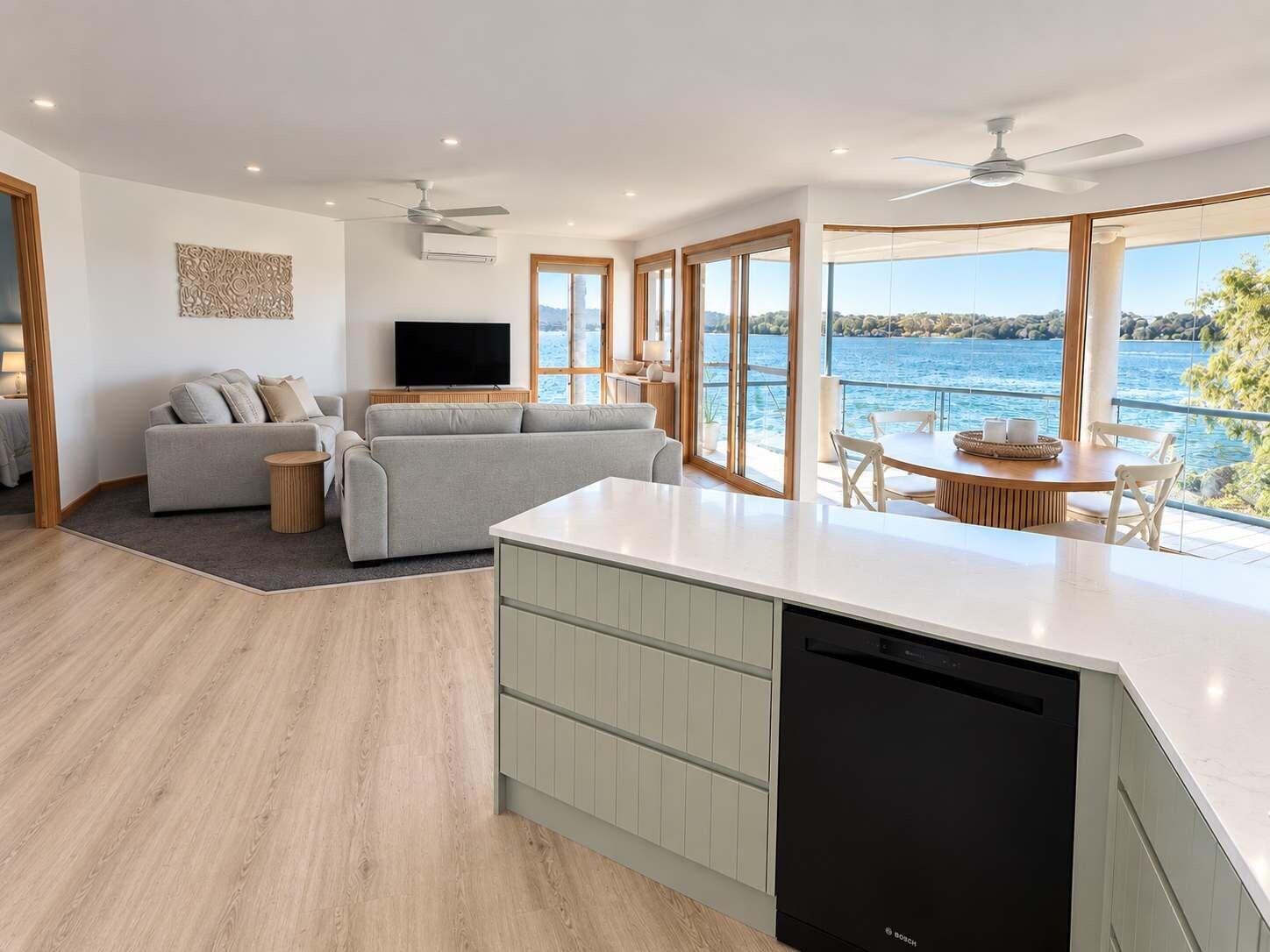A modern kitchen with a Bosch dishwasher, a dining area with a table and chairs, a grey sofa, and a large window overlooking a body of water.