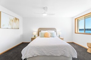 A bedroom with a bed, a ceiling fan, bedside lamps, a window with a view, and a painting on the wall.