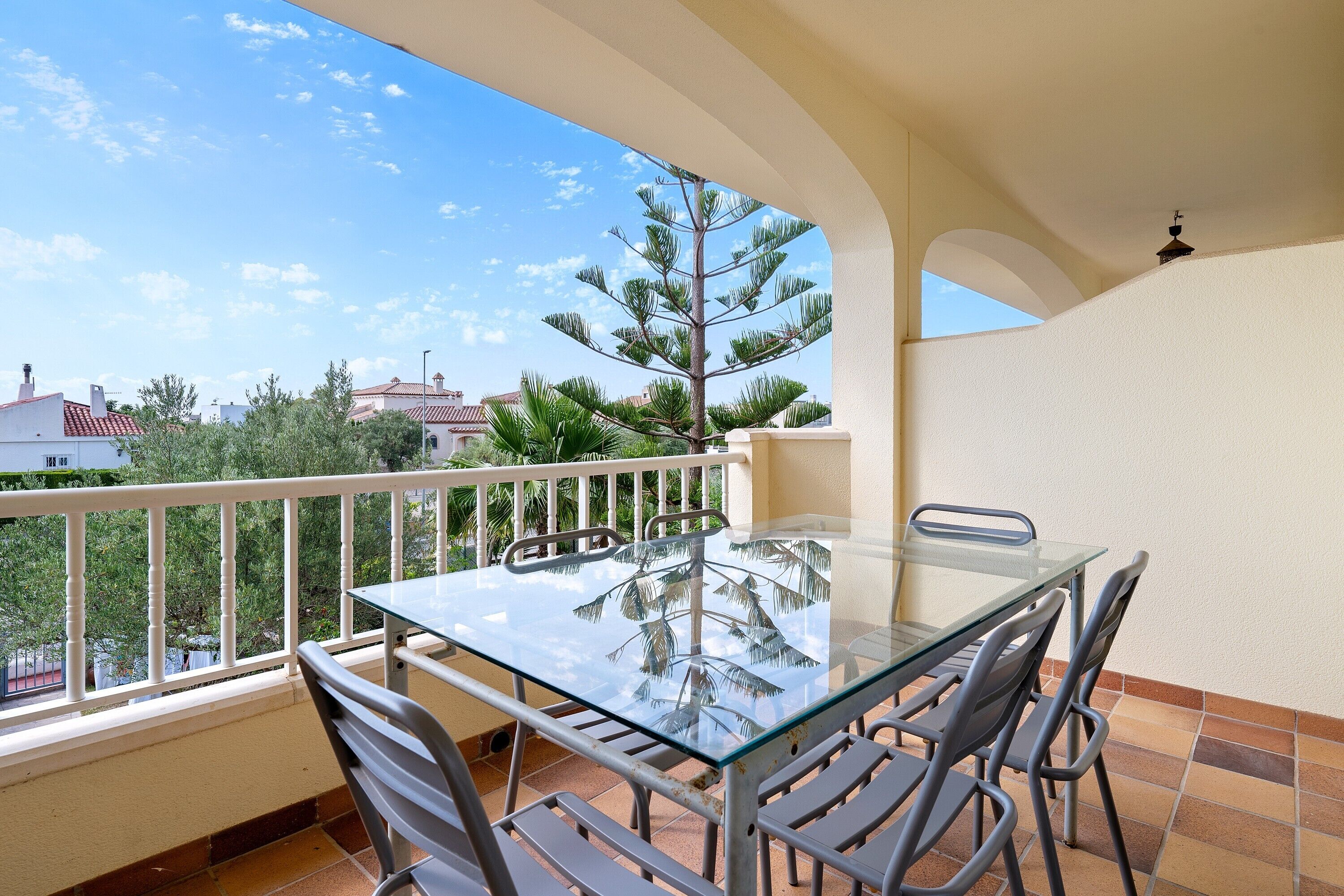A balcony with a glass table and chairs, a white railing, and a view of trees and buildings.