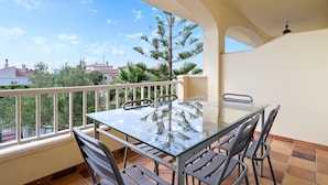 A balcony with a glass table and chairs, a white railing, and a view of trees and buildings.